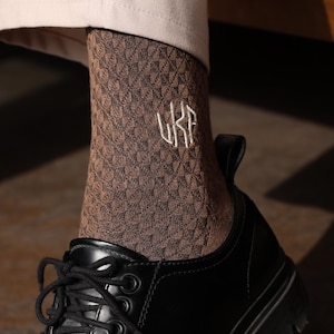 Puede incluir: Primer plano de la parte inferior de la pierna y el pie de una persona. La persona lleva calcetines marrones estampados con las letras "UKR" bordadas en el lateral. La persona también lleva zapatos de cuero negro con suelas gruesas y cordones negros.