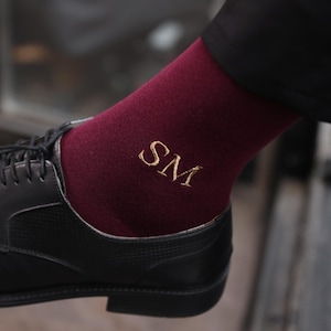 Può includere: Calzini bordeaux con iniziali "SM" ricamate in oro, abbinati a scarpe eleganti in pelle nera. I calzini sono visibili sopra la scarpa, mostrando uno stile classico e personalizzato.
