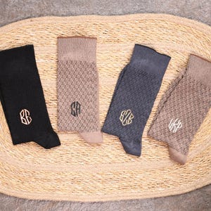 Puede incluir: Cuatro pares de calcetines en negro, beige y gris azulado, cada uno con un monograma. Los calcetines están colocados sobre una alfombra ovalada tejida. Los calcetines tienen un patrón texturizado y son de material suave.