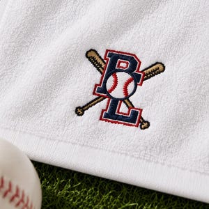 May include: A white terry towelling cloth featuring an embroidered logo of the letters B and L in navy blue with red outlines, positioned over two crossed tan baseball bats and a white baseball with red stitching in the centre of the letters.