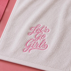 Puede incluir: Una toalla de mano rectangular blanca con el texto bordado en rosa Let's Go Girls en letra cursiva. La toalla tiene una superficie de tela de rizo suave y texturizada con un borde blanco limpio, sobre un fondo de color rosa sólido.