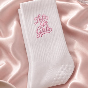 Op de afbeelding: Een witte geribbelde sok met de geborduurde roze tekst Let's Go Girls op het bovenste gedeelte. De sok is voorzien van een reeks kleine, ronde, witte siliconen gripstippen op het hielgedeelte voor extra grip en stabiliteit.