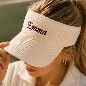 May include: A cream-coloured knitted visor featuring a cable knit texture on the headband and a ribbed brim. The name Emma is embroidered in dark brown script lettering on the front centre of the headband, providing a clear contrast.