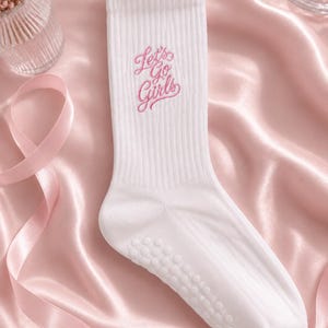May include: A white ribbed crew sock featuring the embroidered pink text Let's Go Girls on the upper shaft. The sole of the sock includes a series of small, circular, white non-slip grip dots for traction, and the fabric has a textured ribbed knit.