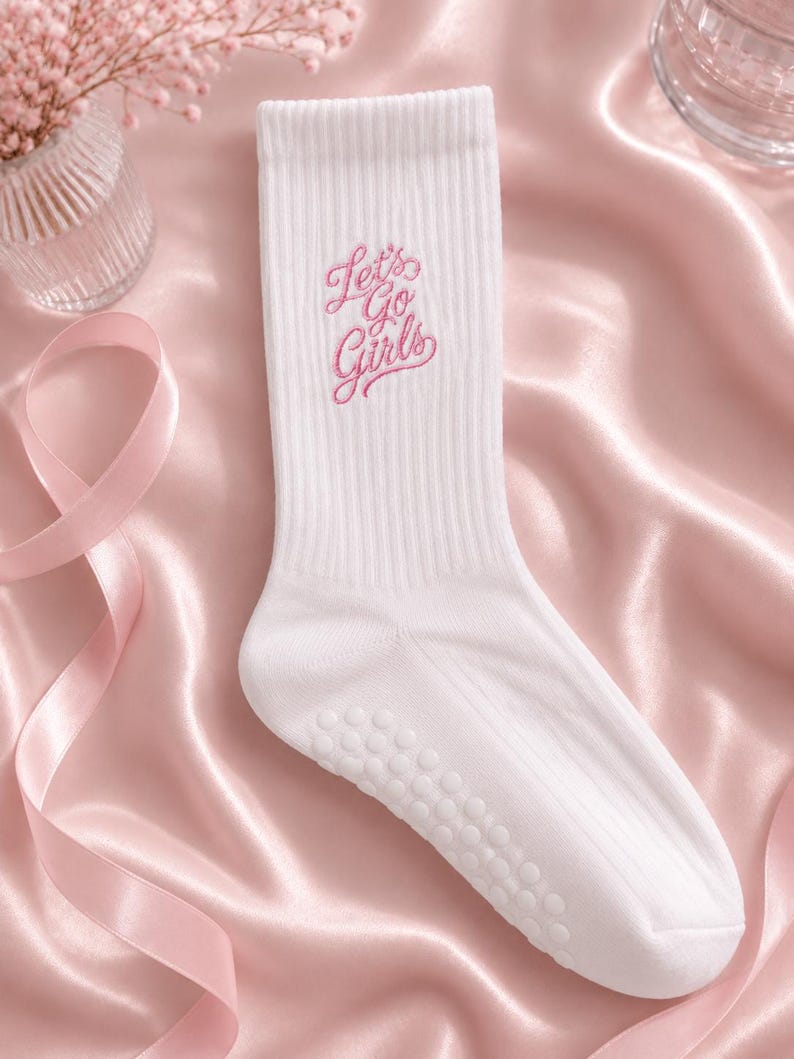 May include: A white ribbed crew sock featuring the embroidered pink text Let's Go Girls on the upper shaft. The sole of the sock includes a series of small, circular, white non-slip grip dots for traction, and the fabric has a textured ribbed knit.