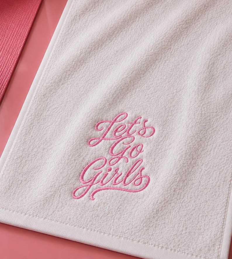 May include: A white rectangular hand towel featuring the embroidered pink cursive text Let's Go Girls. The towel has a soft, textured terry cloth surface and a clean, finished white border edge, presented against a solid pink background.