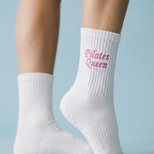 May include: A pair of white crew-length socks featuring ribbed cuffs and the text "Pilates Queen" embroidered in pink script lettering. The socks include small, circular grip dots on the soles to provide traction during physical activity.