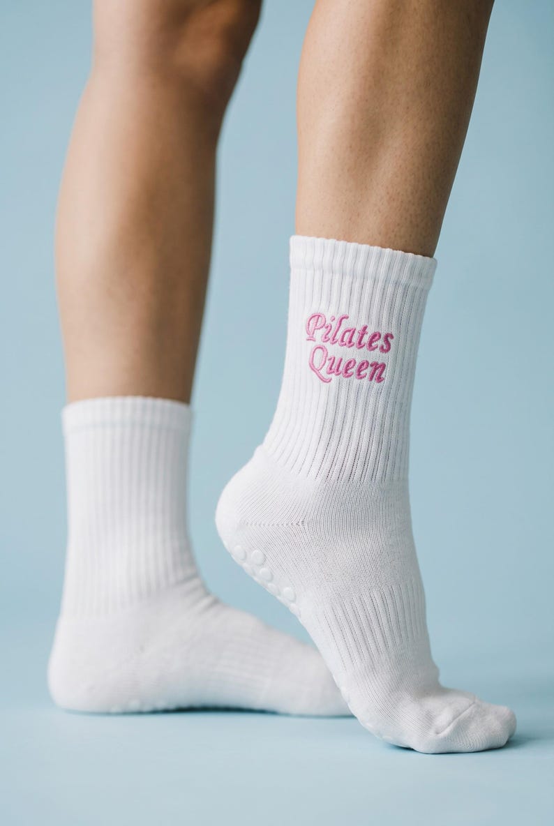 May include: A pair of white crew-length socks featuring ribbed cuffs and the text "Pilates Queen" embroidered in pink script lettering. The socks include small, circular grip dots on the soles to provide traction during physical activity.