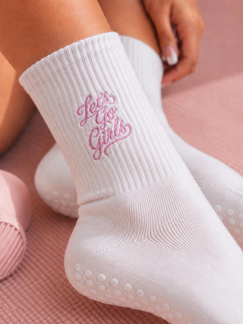 May include: A pair of white crew socks featuring a ribbed texture and the embroidered pink text "Let's Go Girls" on the side. The soles of the socks are equipped with small, white, circular silicone grip dots to provide traction on smooth surfaces.