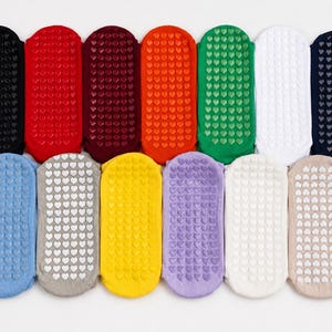 May include: A collection of thirteen low-cut ankle socks arranged in two rows, featuring a variety of solid colors including black, red, maroon, orange, green, white, navy, blue, gray, yellow, purple, cream, and beige with heart-shaped grips.