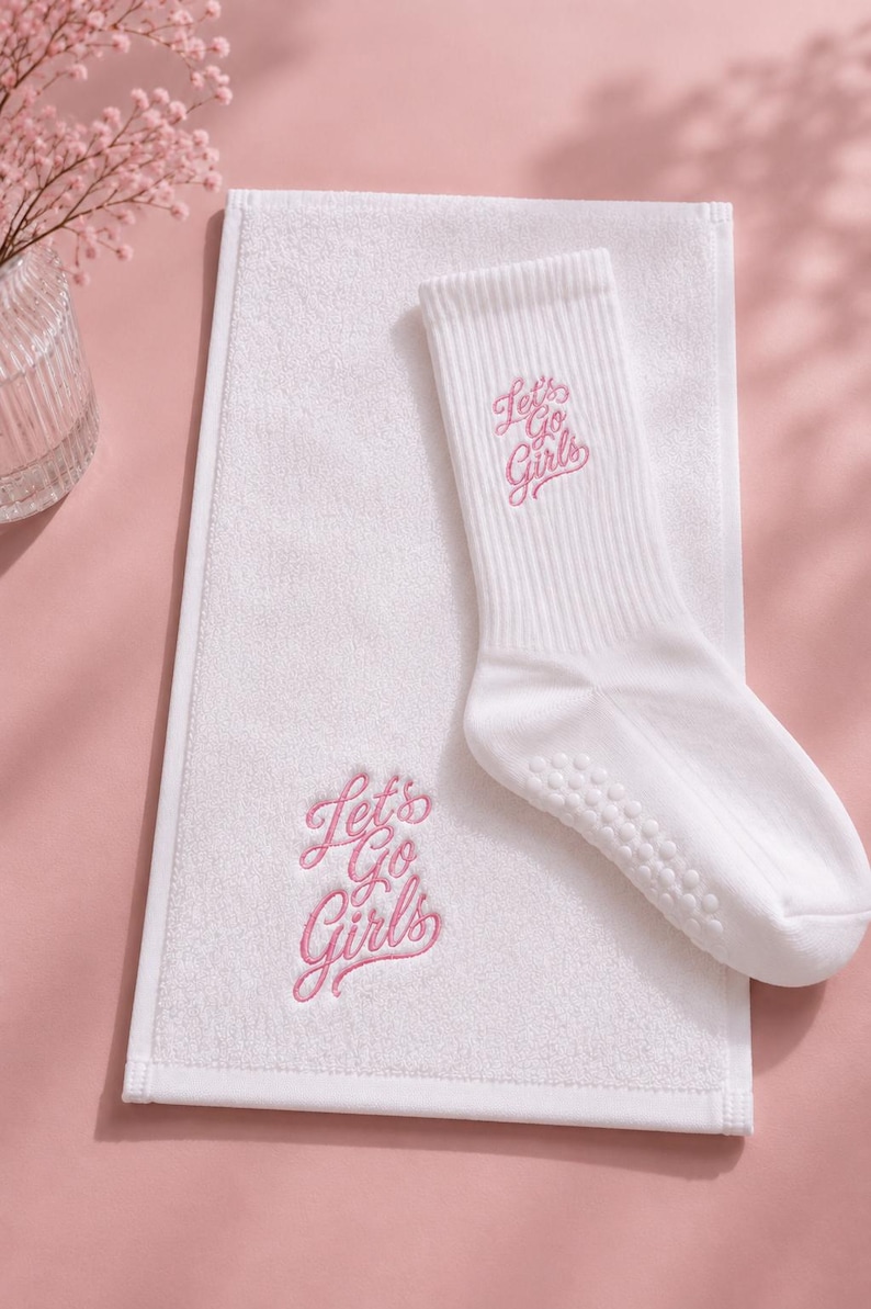 May include: A white rectangular hand towel and a matching white crew sock, both featuring the embroidered pink text Let's Go Girls. The sock has a ribbed cuff and small white silicone grip dots on the sole for traction on smooth surfaces.