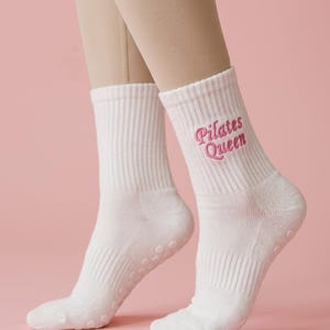 May include: A pair of white crew-length socks featuring the embroidered text "Pilates Queen" in pink thread on the outer side. The socks have a ribbed cuff and small, circular, tonal grip dots on the sole for traction during physical activity.