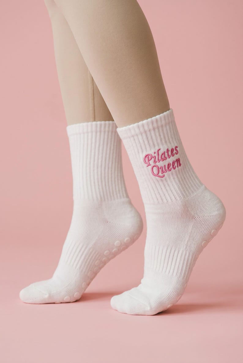 May include: A pair of white crew-length socks featuring the embroidered text "Pilates Queen" in pink thread on the outer side. The socks have a ribbed cuff and small, circular, tonal grip dots on the sole for traction during physical activity.