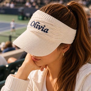 Op de afbeelding: Een crèmekleurige gebreide zonneklep met een geribbelde rand en een hoofdband met kabelpatroon. Op de voorkant van de hoofdband is de naam Olivia geborduurd in donkerblauw cursief schrift, wat een duidelijk contrast vormt met de lichte stof.