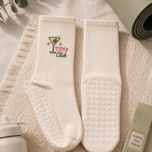 May include: A pair of cream-colored crew socks featuring an embroidered martini glass graphic with an olive and the text "Tipsy Club" in pink and green. The soles of the socks have a repeating pattern of small, raised heart-shaped silicone grips.
