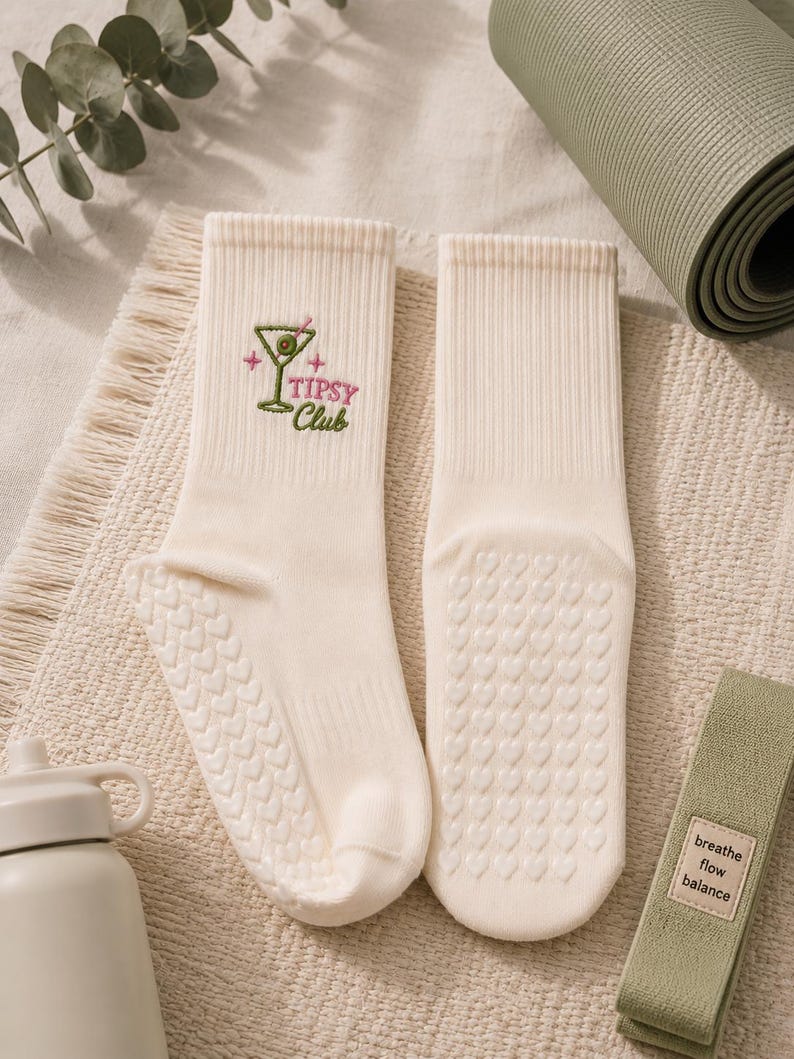 May include: A pair of cream-colored crew socks featuring an embroidered martini glass graphic with an olive and the text "Tipsy Club" in pink and green. The soles of the socks have a repeating pattern of small, raised heart-shaped silicone grips.
