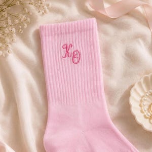 May include: A light pink ribbed crew sock featuring the embroidered letters XO in a darker pink thread. The sock has a vertical ribbed texture throughout the fabric and is positioned against a soft, light-colored fabric background.