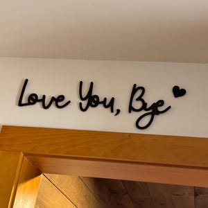 May include: Black wooden wall art with the phrase "Love You, Bye" in a cursive font, accompanied by a small heart symbol. The art is mounted on a white wall above a wooden door frame.