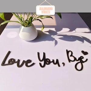 May include: A white, ribbed ceramic vase holds a green plant. Below, the words "Love You, Bye" are spelled out in dark brown, cursive lettering. A small heart shape is next to the word "Bye". A sign that says "HANGING VIBES" is at the top.