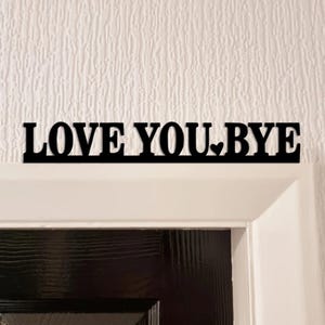 May include: Black metal sign with the words "LOVE YOU. BYE" cut out in a bold font. The word "YOU" has a heart symbol replacing the period. The sign is mounted above a black door.