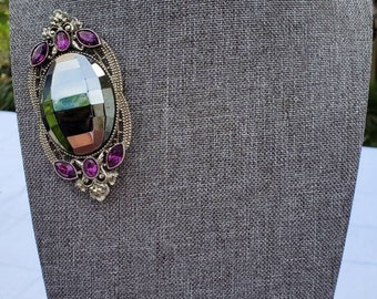 Puede incluir: Un broche de plata con una piedra grande, facetada, de color morado oscuro rodeada de piedras moradas más pequeñas.