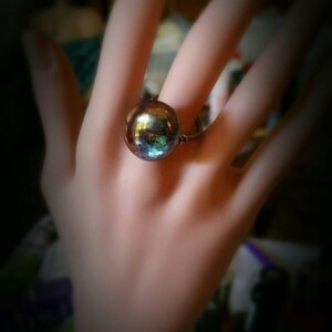 Puede incluir: Un anillo con una gran cuenta redonda de vidrio iridiscente. La cuenta tiene un color azul oscuro, verde y morado.