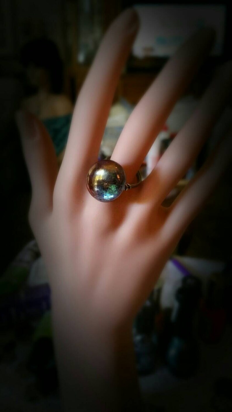 Puede incluir: Un anillo con una gran cuenta redonda de vidrio iridiscente. La cuenta tiene un color azul oscuro, verde y morado.