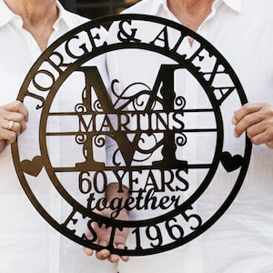 May include: A black metal family sign held by a smiling couple. The sign is circular with the names "Jorge & Alexa" and "Martins" inscribed. It also includes "60 Years together EST 1965" and decorative hearts.