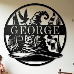 May include: Circular black metal wall art with a wizard hat, castle, scarf, and broom design. The name "GEORGE" is displayed in white. The artwork is mounted on a white wall, suitable for a themed room.