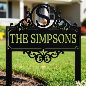 May include: Black metal lawn sign with the name "THE SIMPSONS" in green lettering. The sign features an ornate design with a large letter "S" in a circle at the top. The sign is displayed on a green lawn.