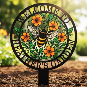 May include: A black metal garden sign with the words "WELCOME TO HEATHER'S GARDEN" in a circular design. The sign features a bee surrounded by orange flowers and green leaves. The sign is placed in the soil.