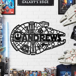 May include: Black metal wall art in the shape of the Millennium Falcon with the name "ANDREWS" cut out in the center. The art piece is mounted on a white brick wall, with Star Wars-themed merchandise in the background.