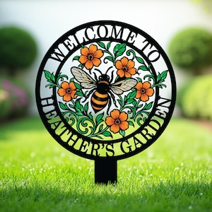 May include: A circular black metal garden stake featuring a cutout design of a central honeybee surrounded by orange flowers and green leaves. The text WELCOME TO HEATHER'S GARDEN is integrated into the circular frame surrounding the floral motif.