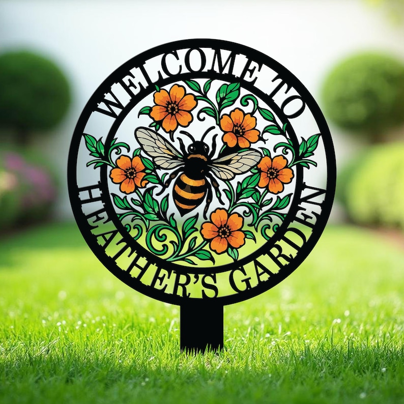 May include: A circular black metal garden stake featuring a cutout design of a central honeybee surrounded by orange flowers and green leaves. The text WELCOME TO HEATHER'S GARDEN is integrated into the circular frame surrounding the floral motif.
