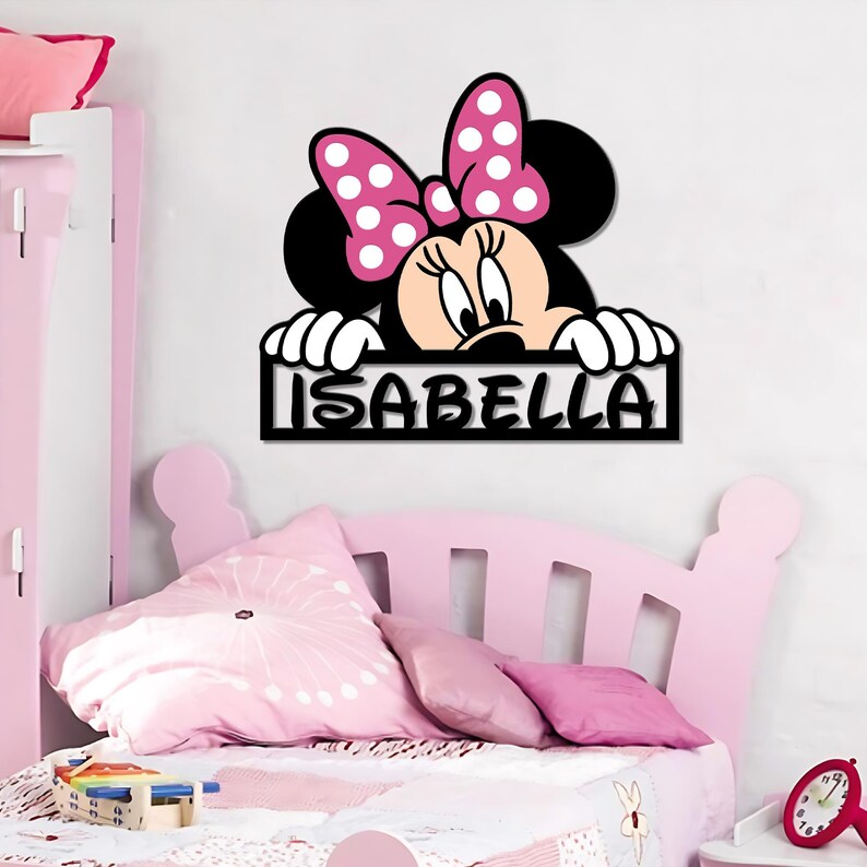 May include: A black wooden sign featuring Minnie Mouse with a pink bow and the name "ISABELLA" in white letters. The sign is designed for a child's bedroom, with a pink bed and pillows in the background.