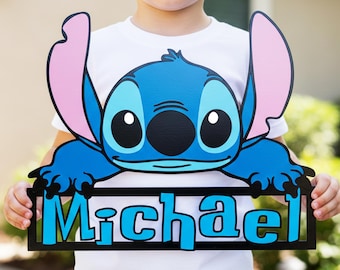 May include: A wooden sign featuring the character Stitch from Lilo & Stitch. The sign is blue, black, and pink, with the name "Michael" in blue letters. The sign is held up by a person wearing a white t-shirt.