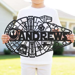 May include: Black metal sign shaped like the Millennium Falcon, with the name "ANDREWS" cut out. The sign incorporates Star Wars elements, such as the Rebel Alliance emblem and Darth Vader's helmet. A decorative item.