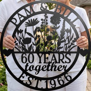 May include: A circular black metal wall sign featuring a floral silhouette design. The text reads PAM & BILL, 60 YEARS together, and EST. 1966. The design includes two small heart shapes on the left and right sides of the circular frame.