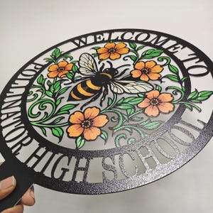 May include: A circular black metal sign featuring a cutout design of a bee surrounded by orange flowers and green leaves. The text "WELCOME TO POLLINATOR HIGH SCHOOL" is cut into the outer border of the circle, which has a textured finish.