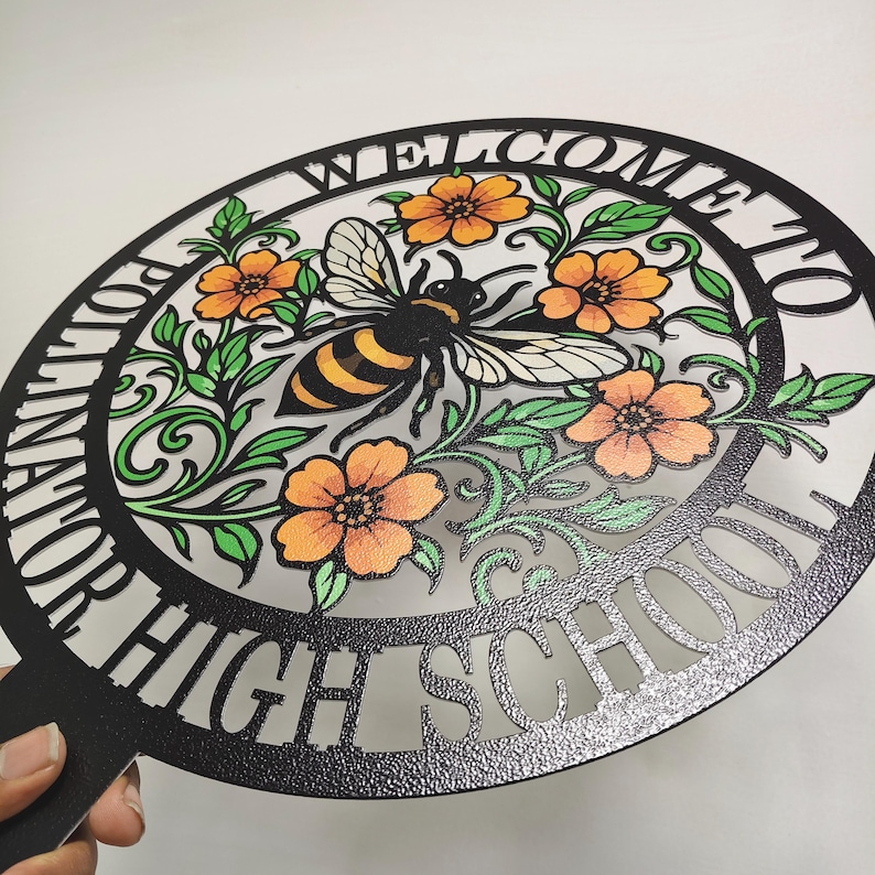 May include: A circular black metal sign featuring a cutout design of a bee surrounded by orange flowers and green leaves. The text "WELCOME TO POLLINATOR HIGH SCHOOL" is cut into the outer border of the circle, which has a textured finish.
