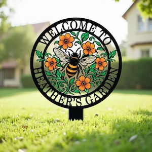 May include: A circular black metal garden stake featuring a cutout design of a bee surrounded by orange flowers and green leaves. The text "WELCOME TO HEATHER'S GARDEN" is integrated into the circular frame, creating a decorative outdoor sign.