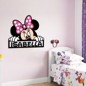 May include: A black Minnie Mouse wall decoration with the name "ISABELLA" in white letters. The character has a pink bow with white polka dots. The bedroom features a bed with a Minnie Mouse-themed comforter and pillowcases.