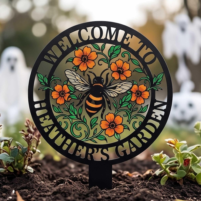May include: A circular black metal garden stake featuring a cutout design of a bee surrounded by orange flowers and green leaves. The text "WELCOME TO HEATHER'S GARDEN" is integrated into the circular border of the sign in a bold, black font.