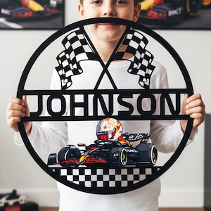May include: Black metal wall art featuring a race car and the name "JOHNSON" in a circular design. Two checkered flags are at the top. The race car is red, black, and yellow. The art is held by a person.