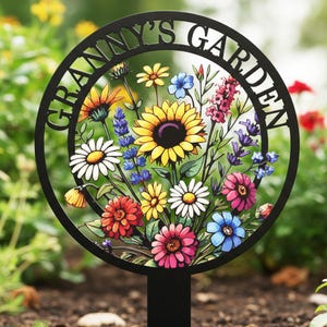 May include: Black metal garden stake with the words "GRANNY'S GARDEN" cut out around a circular frame. The frame contains a colorful arrangement of painted flowers, including sunflowers, daisies, and other blooms. The stake is placed in the soil.