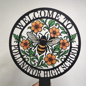May include: A circular black metal sign featuring a central illustration of a bee surrounded by orange flowers and green leaves. The text "WELCOME TO POLLINATOR HIGH SCHOOL" is cut out along the border of the circle. The sign includes a stake at the bottom.