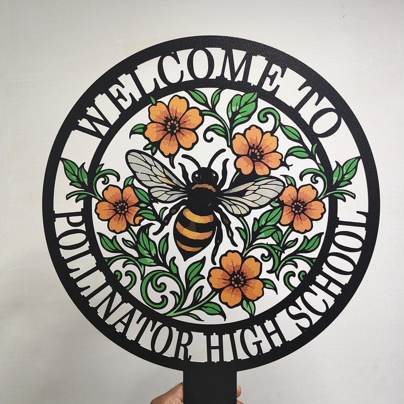 May include: A circular black metal sign featuring a central illustration of a bee surrounded by orange flowers and green leaves. The text "WELCOME TO POLLINATOR HIGH SCHOOL" is cut out along the border of the circle. The sign includes a stake at the bottom.