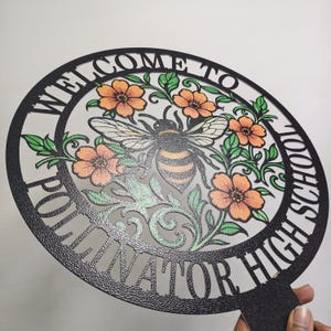 May include: A circular black metal sign with a textured finish featuring the text "WELCOME TO POLLINATOR HIGH SCHOOL" along the border. The center displays a detailed illustration of a bee surrounded by orange flowers and green leaves.