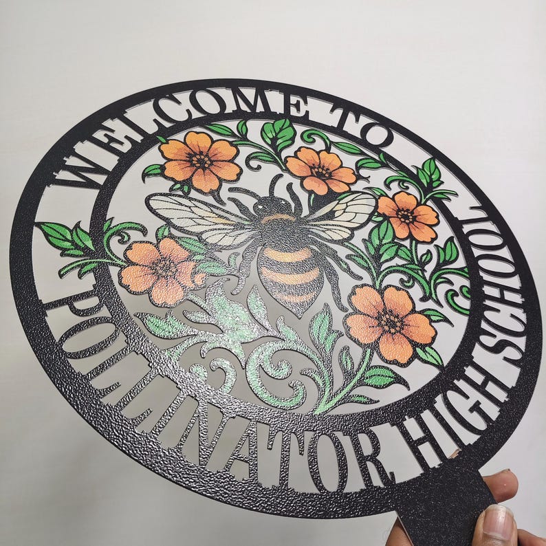 May include: A circular black metal sign with a textured finish featuring the text "WELCOME TO POLLINATOR HIGH SCHOOL" along the border. The center displays a detailed illustration of a bee surrounded by orange flowers and green leaves.