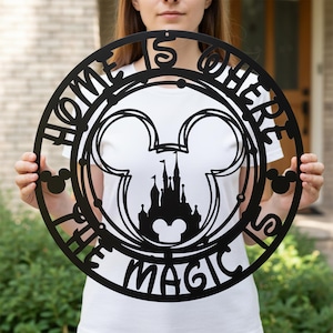 以下が含まれることがあります： 黒い金属製のウォールアート。円形のデザインで、「HOME IS WHERE THE MAGIC IS」の文字が入っています。ミッキーマウスの耳と城のシルエットが含まれています。家の装飾用の装飾品です。
